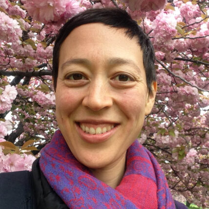 Alison Lin, an East Asian person, smiles in front of a cherry blossom tree. She wears a pink and purple scarf and smiles at the camera