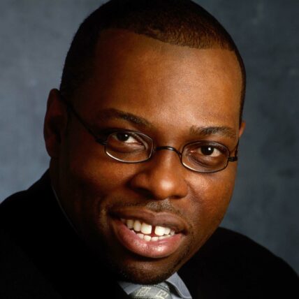 David Greene III, a Black person, wears glasses and a suit, smiling at the camera