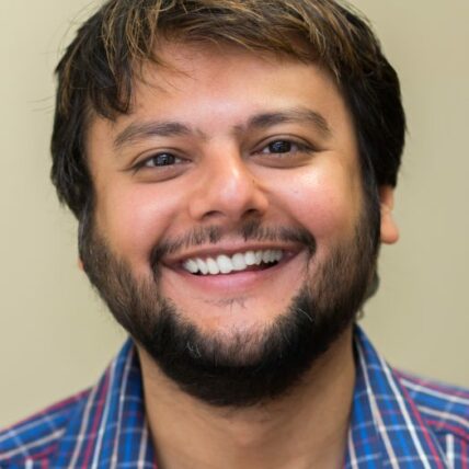 Zulayka Santiago wears a blue, red, and white checkered shirt and has a short beard, smiling at the camera