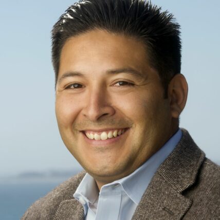 Vince Marquez, smiles at the camera, wearing a blue button up and brown tweed blazer