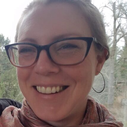 Headshot of Chandra Larsen, wearing glasses, hoop earrings and a brown scarf. Their hair is back and they are smiling at the camera