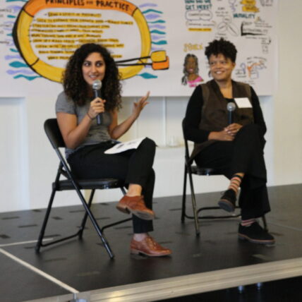 Natalie Bamdad and Elissa Sloan Perry facilitating, both seated in front of graphic recordings, speaking into microphones