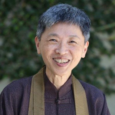 Photo of Norma Ryuko Kaweloku Wong Roshi, with short cropped salt and pepper hair, smiling at the camera wearing a dark purple top and olive green sash against a blurred background of foliage.