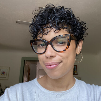 Photo of Naima Yael Tokunow, a Black woman with short curly black hair, wears tortoiseshell cat-eye glasses, wavy gold hoops and a white top, smiles at the camera.