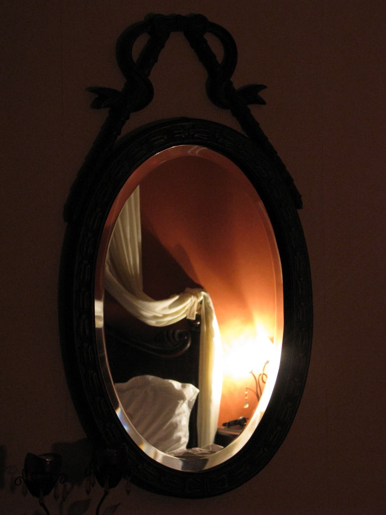 A photograph of a mirror reflecting a bed at night, a soft yellow glow shines from a bedside lamp.